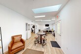 Located at 2180 Hawthorn Meadows in Howell, Michigan, this single-floor amenity space offers a well-appointed and versatile environment designed for both relaxation and activity. The interior is characterized by crisp white walls, light wood-style flooring, and an abundance of natural light streaming in through prominent skylights and large windows — creating an open, airy atmosphere throughout.

The main gathering area serves as the heart of the space, featuring a mix of high-top bistro tables with industrial-style stools, a comfortable lounge sofa, and a dining table with chairs. A wall-mounted TV adds to the functionality of the room, while views of surrounding greenery through the windows enhance the inviting ambiance.

The kitchen is sleek and modern, outfitted with white shaker-style cabinetry, bright white countertops, a stainless steel refrigerator, a dishwasher, microwave, and a peninsula with bar seating — ideal for casual gatherings or community events.

A dedicated fitness room rounds out the space, equipped with treadmills, a full dumbbell rack, a stability ball, and additional cardio equipment. A floor-to-ceiling mirror amplifies the room, and blue lockers provide convenient storage for residents. The fitness area is well-lit and neatly organized, making it a practical and motivating workout environment.