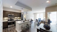 This single-floor residence showcases a thoughtfully designed open-concept layout that connects the kitchen, dining, and living areas into one cohesive, flowing space. The kitchen is a standout feature, appointed with rich dark cabinetry, stainless steel appliances, a textured tile backsplash, and light quartz countertops that extend to a generously sized island with bar seating — ideal for casual dining or entertaining. Stylish pendant lighting with clear glass fixtures hangs above the island, adding a contemporary touch. The kitchen transitions seamlessly into the dining area, where a dark wood table provides a natural contrast to the lighter surroundings. The living area is bright and airy, benefiting from large windows dressed in sheer white curtains that allow abundant natural light to fill the room. Wide-plank hardwood-style flooring runs throughout, grounding the space with warmth and continuity. The living room is furnished with a neutral sofa, a classic blue accent chair, and a simple coffee table, creating a comfortable and inviting atmosphere. Soft, neutral wall tones and clean architectural lines give the entire space a modern yet welcoming character.