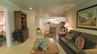 This single-floor residence offers a well-appointed and cohesive living environment designed for both comfort and functionality. The living room serves as the heart of the space, featuring plush carpeted flooring, warm neutral-toned walls, and crown molding that adds a refined architectural touch. Recessed lighting illuminates the room alongside warm ambient lamps, creating a cozy and welcoming atmosphere. The living area is thoughtfully arranged with a cushioned sofa accented with decorative pillows, a classic upholstered armchair, and a rich wood coffee table adorned with lush greenery, lending a homey and lived-in feel. A substantial wood hutch along one wall provides ample display and storage space. The open-concept layout connects the living room directly to the kitchen through a pass-through counter, promoting an easy flow between spaces. The kitchen is outfitted with white cabinetry, full-sized appliances including a refrigerator and stove, and a clean countertop workspace. Potted plants lining the top of the kitchen cabinetry bring a touch of nature indoors. A small decorative accent table transitions the space into what appears to be a secondary area or hallway, further enhancing the functional layout of this inviting residence.