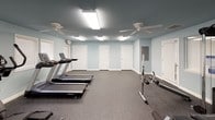 This fitness center is a thoughtfully designed single-floor exercise space finished in soft blue-gray tones that lend a calm, focused atmosphere. The durable speckled rubber flooring provides cushioning and traction throughout, making it well-suited for a variety of workout activities. Overhead, multiple fluorescent light fixtures ensure consistent, bright illumination across the entire room, supplemented by two ceiling fans that promote air circulation during intense sessions. Several windows fitted with horizontal blinds allow natural light to filter in while maintaining privacy.

The space is comprehensively outfitted with a range of exercise equipment. Cardio options include multiple treadmills and an elliptical trainer, while strength training is supported by a multi-station cable machine, a weight bench, a rowing machine, and free weights. A wall-mounted water fountain provides convenient hydration access. Multiple white interior doors offer separate entry and exit points, and an illuminated exit sign underscores the room's compliance with safety standards. A wall clock allows users to monitor their workout duration. The combination of ample equipment variety, practical amenities, and a clean, organized layout makes this fitness center a functional and welcoming environment for exercise.
