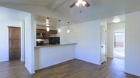 This single-floor space offers an open-concept design that seamlessly connects the living and kitchen areas, creating a cohesive and functional layout. The kitchen is defined by rich wood cabinetry, granite countertops, and black appliances — including a range and refrigerator — all centered around a raised peninsula that doubles as a breakfast bar. Three elegant pendant lights hang above the peninsula, providing focused task lighting while enhancing the overall aesthetic. The living area features warm, dark hardwood flooring that extends throughout, grounding the space with a sense of richness and continuity. A ceiling fan with a light fixture is mounted above, complementing the exposed wooden ceiling beams that add architectural character and a touch of rustic charm. The clean white walls throughout create a bright, neutral backdrop that highlights the natural tones of the wood elements. A hallway off the main living area suggests additional rooms beyond, offering practical separation within the layout. Overall, the space strikes a balance between everyday functionality and inviting warmth, making it well-suited for comfortable living.