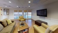 Located at 1 River Court in Amesbury, Massachusetts, this single-floor space is thoughtfully designed to offer a warm, functional, and inviting environment. The layout flows seamlessly across several distinct areas, each serving a specific purpose while maintaining a cohesive aesthetic throughout.

The main lounge area is anchored by comfortable upholstered seating, warm hardwood flooring, and a large flat-screen television mounted on the wall. Multiple windows dressed with woven Roman shades allow generous natural light to filter in, creating a bright and relaxed atmosphere. A recessed-lit drop ceiling adds a polished, finished look.

A second seating area features a classic fireplace with a decorative mantel, providing a cozy focal point. This room is furnished with plush sofas and armchairs arranged around a central coffee table, ideal for casual gathering or quiet relaxation. Wainscoting along the lower walls adds a refined architectural detail throughout the space.

The kitchenette is well-equipped with warm maple cabinetry, dark granite countertops, a stainless steel microwave, and a compact refrigerator — offering convenient amenities for everyday use.

A bright, windowed workspace provides a dedicated area for productivity, furnished with a desk, seating, and computer equipment, surrounded by large windows that frame outdoor views of the natural landscape. Hardwood flooring and recessed lighting carry through this area as well, ensuring visual continuity across the entire space.