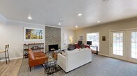This well-appointed single-floor community space is designed to offer residents a comfortable and functional gathering environment. The centerpiece of the main lounge area is a striking floor-to-ceiling stacked stone fireplace, which anchors the room with warmth and visual interest. The lounge is furnished with a neutral sofa, cognac leather accent chairs, and layered rugs over plush carpeting, creating an inviting atmosphere ideal for relaxing or socializing. Recessed lighting and crown molding throughout add a clean, polished finish to the space.

Adjacent to the lounge, a dedicated workspace features multiple computer stations positioned near a large window that fills the area with natural light. A functional kitchen offers white cabinetry, dark granite countertops, a mosaic tile backsplash, black appliances, and a peninsula bar with seating — ideal for casual dining or entertaining. The hardwood-style flooring in the kitchen provides a warm contrast to the surrounding carpeted areas.

A separate dining nook with a wooden table and modern wire-frame chairs opens directly onto the pool area through full-length French doors, flooding the space with natural light and offering seamless indoor-outdoor connectivity. Views of the fitness area through interior windows further underscore the multi-purpose nature of this thoughtfully designed community hub.