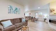 This well-designed single-floor residence offers a clean, contemporary aesthetic throughout, characterized by crisp white walls, light-toned luxury vinyl plank flooring, and an abundance of natural light from multiple windows. The open-concept layout seamlessly connects the living room, dining area, and kitchen, creating a spacious and cohesive living environment. The living room provides a comfortable gathering space, while the adjacent dining area features a round table with modern wishbone chairs and direct access to an exterior door. The kitchen is thoughtfully appointed with dark cabinetry, stainless steel appliances, white countertops, and a peninsula breakfast bar with seating — ideal for casual dining. A wall-mounted mini-split system ensures efficient climate control throughout the main living space. The residence includes two carpeted bedrooms, each benefiting from natural light through well-positioned windows and closet storage. The bathroom features a vanity with white countertops and dark cabinetry, consistent with the home's design language. A dedicated in-unit laundry closet with a full-size washer and dryer adds significant convenience. Overall, the space presents a modern, functional, and inviting atmosphere suited to comfortable everyday living.