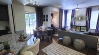 This single-floor space showcases a thoughtfully designed open-concept layout where the dining and living areas flow seamlessly into one another, creating a cohesive and inviting atmosphere. Rich hardwood flooring runs throughout, anchoring the space with warmth and elegance.

The dining area features a sleek round table surrounded by contemporary dark chairs, positioned to take full advantage of the natural light streaming in through large sliding glass doors that open to a lush outdoor view. Dramatic floor-to-ceiling curtains in deep purple add a bold, sophisticated accent against the neutral walls.

The living area is generously proportioned, centered around a plush sofa and complemented by stylish ottoman seating. A patterned area rug defines the lounge zone, while recessed lighting and track lighting provide layered illumination throughout. Decorative elements such as framed wall art, accent lamps, and carefully chosen furnishings contribute to the space's polished, contemporary aesthetic.

Large windows on multiple walls flood the interior with natural light, enhancing the open and airy feel of the space. The overall ambiance is modern and refined, with a curated color palette of neutral tones punctuated by rich purples and greens, creating a space that is both functional and visually striking.