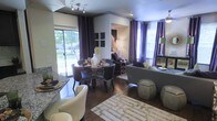 This single-floor space showcases a thoughtfully designed open-concept layout where the dining and living areas flow seamlessly into one another, creating a cohesive and inviting atmosphere. Rich hardwood flooring runs throughout, anchoring the space with warmth and elegance.

The dining area features a sleek round table surrounded by contemporary dark chairs, positioned to take full advantage of the natural light streaming in through large sliding glass doors that open to a lush outdoor view. Dramatic floor-to-ceiling curtains in deep purple add a bold, sophisticated accent against the neutral walls.

The living area is generously proportioned, centered around a plush sofa and complemented by stylish ottoman seating. A patterned area rug defines the lounge zone, while recessed lighting and track lighting provide layered illumination throughout. Decorative elements such as framed wall art, accent lamps, and carefully chosen furnishings contribute to the space's polished, contemporary aesthetic.

Large windows on multiple walls flood the interior with natural light, enhancing the open and airy feel of the space. The overall ambiance is modern and refined, with a curated color palette of neutral tones punctuated by rich purples and greens, creating a space that is both functional and visually striking.