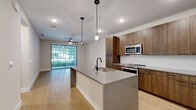 This single-floor residence showcases a cohesive modern design with clean lines and contemporary finishes throughout. The open-concept kitchen serves as a central highlight, anchored by a spacious island topped with grey quartz countertops and fitted with a matte black faucet and undermount sink. Warm-toned wood cabinetry lines the walls from floor to ceiling, complementing the stainless steel appliances, including a gas range, microwave, and refrigerator. Globe pendant lights suspended above the island add a stylish, curated touch, while light wood-tone flooring extends seamlessly into the adjacent living area.

The living space is bright and open, benefiting from a large sliding glass door with vertical blinds that frames outdoor greenery and allows natural light to fill the room. A ceiling fan overhead provides practical comfort.

The bedrooms are finished with plush neutral carpet and feature generous closet space, including walk-in configurations. Large windows with exterior views and ceiling fans contribute to an airy feel.

The bathroom presents a refined aesthetic with a floating wood vanity, quartz countertop, matte black hardware, and a large backlit mirror that lends a clean, contemporary glow to the space. Neutral tile flooring completes the polished look.