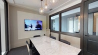 This single-floor office space centers around a well-appointed conference room designed for professional meetings and presentations. The focal point is a long marble-topped conference table with distinctive white and grey veining, surrounded by ergonomic dark chairs and equipped with built-in cable management for seamless connectivity.

Overhead, a cluster of globe-shaped glass pendant lights suspended from brass fixtures provides warm, ambient illumination that complements the neutral tones of the walls and tray ceiling. A large wall-mounted flat-screen display is positioned prominently at one end of the room, ideal for presentations and video conferencing, while a frosted glass whiteboard panel on the opposite wall offers an additional tool for collaboration.

The room is enclosed by floor-to-ceiling glass partitions with sleek dark frames, offering visual transparency to adjacent spaces while maintaining acoustic separation. Beyond the glass walls, a warm-toned wood slatted shelving unit and sheer white drapery are visible, contributing to the refined aesthetic. A dark-framed glass door provides access from the corridor, where textured carpet flooring continues throughout. The overall design balances functionality with a polished, contemporary aesthetic suited to a professional office environment.