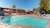 Located at 7761 Greenback Ln in Citrus Heights, California, this three-floor residential community offers a well-appointed outdoor amenity area designed for relaxation and social gathering. The centerpiece of the outdoor space is a generously sized swimming pool with clear, blue-green water, framed by a wide concrete sun deck furnished with wicker chaise lounges and vibrant red and blue patio umbrellas. Adjacent to the pool, a dedicated spa provides an additional retreat for residents.

A standout feature of the community is the covered outdoor pavilion, constructed with striking exposed wood beam rafters and stone accent columns. This structure houses a fully equipped outdoor kitchen and dining area, offering a comfortable and shaded space for entertaining. The pavilion's architectural detail adds a refined, lodge-inspired aesthetic to the overall outdoor environment.

The surrounding landscape features mature redwood and deciduous trees that provide natural shade and a sense of seclusion, enclosed by decorative iron fencing. A dedicated pool house with restroom facilities serves the outdoor area. The multi-story residential buildings feature neutral siding and pitched rooflines that complement the natural surroundings. Under California's clear blue skies, the community presents a cohesive and inviting outdoor living environment throughout.