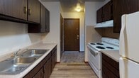 Located at 1223 Oak Hill Ct in Toledo, Ohio, this single-floor unit offers a practical and well-organized layout suited for comfortable everyday living. The kitchen is efficiently designed with dark wood cabinetry on both sides, a double-basin stainless steel sink, laminate countertops, a white electric range with overhead ventilation, and durable hardwood-style flooring. A ceiling fan with a light fixture illuminates the space, and a solid wood entry door provides direct access from the kitchen area. The living area is generously sized, featuring plush wall-to-wall carpet, neutral beige walls, and a sliding glass door dressed with vertical blinds that invites natural light and provides outdoor access. A wall-mounted air conditioning unit serves the space for climate comfort. The carpeted bedrooms are well-proportioned, each offering windows with blinds that bring in natural light, baseboard heating units, and closet access. The full bathroom is cleanly appointed with a soaking tub and shower combination, a toilet, wood-framed vanity with a white countertop sink, and a lighted mirror cabinet above. Overall, the unit presents a clean, functional aesthetic with consistent neutral tones, carpet in living and sleeping areas, and hard flooring in the kitchen throughout its single level.