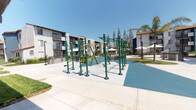 Located in Chatsworth, Los Angeles, this outdoor community amenity space is designed to support an active and social lifestyle. The centerpiece of the area is a well-equipped outdoor fitness station, featuring a variety of green powder-coated exercise apparatus including pull-up bars, resistance cable machines, balance elements, and stretching stations, all set atop a rubberized safety surface. The fitness zone is complemented by a dedicated BBQ and outdoor kitchen area, complete with built-in grills and counter space, alongside a shaded dining setup with an umbrella-covered table, ideal for outdoor gatherings. Lush, manicured green lawns border the concrete pathways that wind through the courtyard, and mature trees including palms and evergreens provide natural shade and greenery. The surrounding residential buildings display a classic Spanish-inspired architectural style, characterized by stucco exteriors, terracotta tile rooflines, and private balconies. String lights are strung between poles, adding a welcoming ambiance for evening use. Glass entry doors with sleek metal framing provide access to the complex. The entire space benefits from abundant Southern California sunshine, creating a bright, open-air environment that encourages recreation and community connection year-round.