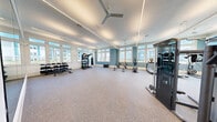 This generously sized fitness center offers a comprehensive workout facility spread across two distinct exercise rooms, delivering an impressive range of training options in a bright and inviting environment. The main exercise room is a standout space, featuring high ceilings with sleek linear lighting, multiple ceiling fans, and an expansive bank of windows that flood the interior with natural light and offer views of the surrounding area. Accent walls in a calming teal-blue complement the clean white trim and speckled rubber flooring, creating an energizing yet comfortable atmosphere. This room is thoroughly equipped with free weights including a full dumbbell rack, barbells, weight plates, a power rack, cable machines, and cardio equipment such as treadmills and rowing machines. A second, more intimate exercise room provides a focused studio environment with light hardwood flooring, additional ceiling fans, and generous window views. It is outfitted with spin bikes, medicine balls, resistance equipment, and open floor space ideal for functional training or group exercise. Built-in shelving and glass-paneled doors connect the two rooms seamlessly, enhancing both flow and visibility throughout the facility. Together, the two rooms form a well-rounded fitness destination suited for a wide variety of training styles and fitness goals.