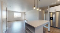 This single-floor residence offers a thoughtfully designed open-concept layout that seamlessly connects the kitchen, dining, and living areas. The kitchen is a standout feature, equipped with stainless steel appliances including a French door refrigerator, electric range, microwave, and dishwasher. White shaker-style cabinetry pairs elegantly with light quartz countertops and a hexagonal tile backsplash, while a central island with built-in shelving provides additional prep space and storage. Stylish globe pendant lights hang above, adding a modern touch. Warm-toned hardwood-style flooring runs throughout the main living areas, enhancing the bright, airy atmosphere created by large windows that invite generous natural light. The spacious living area offers direct access to an outdoor space through a glass door. Bedrooms are finished with plush neutral carpet, providing a comfortable retreat. The primary bathroom is well-appointed with a dual-sink vanity featuring illuminated mirrors, ample built-in shelving, and a tub-shower combination adorned with marble-look tile surround. The overall design is clean, contemporary, and move-in ready, with a neutral color palette that complements a wide range of décor styles.