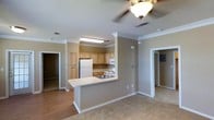 This single-story space showcases an open-concept layout that seamlessly connects the kitchen and living areas. The kitchen is equipped with light wood cabinetry, white appliances including a refrigerator, electric range, and microwave, complemented by a peninsula with a light-colored countertop that doubles as a breakfast bar. A stainless steel sink is integrated into the peninsula, enhancing both functionality and flow. The flooring transitions from hardwood-style material in the kitchen to carpeting in the living area, adding textural contrast throughout. Warm neutral wall tones are carried consistently across the space, contributing to a cohesive aesthetic. Crown molding runs along the ceiling perimeter, lending a polished, finished look, while a ceiling fan with a light fixture provides both comfort and illumination. Doorways visible from the main living area suggest additional rooms beyond, including what appears to be bedrooms and a bathroom. The layout offers a practical, well-organized design suitable for comfortable everyday living.