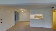 This single-floor residence offers an open-concept design that seamlessly connects the living area and kitchen. The living space is finished with wall-to-wall carpeting in a neutral beige tone, contributing to a warm and understated ambiance. A modern track light fixture is mounted to the ceiling, providing direct illumination across the room. Vertical blinds cover a sliding glass door on the left wall, offering access to an outdoor area while allowing for light control and privacy.

The kitchen is positioned at the far right and is defined by a raised peninsula counter that creates a natural divide between the cooking and living spaces. White cabinetry lines the upper walls, and white appliances — including a microwave and a gas range with a built-in sink — are neatly integrated into the layout. The countertops are light-toned, maintaining the cohesive, clean aesthetic found throughout the unit.

Textured ceilings and a structural beam detail add modest architectural character to the space. The overall palette is light and neutral, creating a practical and functional environment.