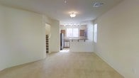 This two-story residence offers a well-organized layout designed for comfortable everyday living. The main floor centers around an open-concept living and kitchen area, where soft carpeting in the living space transitions toward the kitchen zone. The kitchen is equipped with stainless steel appliances, including a refrigerator and dishwasher, and features warm wood-toned cabinetry paired with a breakfast bar that provides both additional prep space and casual seating. Track lighting above the kitchen ensures the area is well-lit, while a flush-mount ceiling fixture illuminates the living area. Natural light filters in through multiple windows fitted with blinds, brightening the neutral, cream-toned walls that give the space a clean and inviting atmosphere. A visible staircase near the entry leads to the second floor, suggesting additional living or sleeping quarters above. The overall design emphasizes an efficient, functional floor plan with a cohesive, understated aesthetic that makes the space easy to personalize.