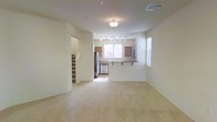 This two-story residence offers a well-organized layout designed for comfortable everyday living. The main floor centers around an open-concept living and kitchen area, where soft carpeting in the living space transitions toward the kitchen zone. The kitchen is equipped with stainless steel appliances, including a refrigerator and dishwasher, and features warm wood-toned cabinetry paired with a breakfast bar that provides both additional prep space and casual seating. Track lighting above the kitchen ensures the area is well-lit, while a flush-mount ceiling fixture illuminates the living area. Natural light filters in through multiple windows fitted with blinds, brightening the neutral, cream-toned walls that give the space a clean and inviting atmosphere. A visible staircase near the entry leads to the second floor, suggesting additional living or sleeping quarters above. The overall design emphasizes an efficient, functional floor plan with a cohesive, understated aesthetic that makes the space easy to personalize.