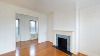This single-floor space showcases a blend of classic architectural charm and clean, minimalist design. The main living area is anchored by a decorative fireplace framed by an elegant white mantel with ornate molding details and a striking blue mosaic tile surround, adding a distinctive focal point to the room. The hearth features a black slate base, complementing the overall aesthetic.

Warm-toned hardwood floors run throughout the space, lending a sense of continuity and natural warmth. The white walls and white ceiling create a bright, open atmosphere, allowing natural light to play across the surfaces. A doorway leads to an adjoining room, which receives natural light through a window overlooking the exterior. A cast-iron radiator is visible in this secondary room, indicative of a traditional heating system consistent with classic building construction.

The space maintains a neutral, versatile palette that provides a clean canvas. Architectural details such as the window and door moldings, along with the fireplace mantel, reflect a period-influenced design sensibility that adds character and depth to the otherwise understated interior.