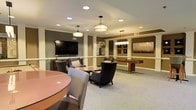 This single-floor community lounge space is thoughtfully designed to offer both relaxation and recreation in a refined setting. The room is anchored by a large wall-mounted flat-screen television, positioned above a low media console and framed by warm, neutral-toned walls accented with classic white wainscoting and decorative columns that lend a sense of architectural elegance.

Seating is arranged to encourage socializing, with a grouping of upholstered armchairs and accent tables positioned in front of the TV, ideal for casual gatherings or quiet evenings. A round table with additional seating is featured prominently in the foreground, offering a comfortable spot for small group activities.

A dedicated games area is visible in the background, highlighted by a full-length shuffleboard table set beneath a distinctive multi-light pendant fixture, adding a recreational focal point to the space. Artwork adorns the walls, contributing to the warm and curated ambiance of the room.

Lighting plays a key role in the overall atmosphere, with a mix of recessed ceiling lights and stylish hanging pendant fixtures casting a warm, inviting glow throughout. Neutral carpeting ties the space together, complementing the earthy tones of the walls and furnishings for a cohesive, welcoming environment suited for both socializing and leisure.