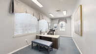 Located at 600 River Birch Ct in Clermont, FL, this single-floor professional office space offers a well-appointed and functional layout designed to support a productive work environment. The space is thoughtfully organized across several distinct work areas, each finished with soft blue-grey carpeting and neutral grey walls that create a calm, professional atmosphere throughout.

The primary workstation area features an L-shaped desk with built-in storage, accompanied by a padded bench seat — well-suited for client consultations or collaborative work. Multiple windows dressed with decorative valances allow generous natural light to fill the room, while recessed and flush-mount fluorescent lighting ensures consistent illumination. Framed artwork adorns the walls, adding a polished, welcoming touch.

A secondary office area, accessible through French doors, provides a private workspace with its own desk and computer setup, along with a large statement painting that lends warmth to the room. An additional area accommodates office equipment, including a multifunction printer, with built-in cabinetry providing ample storage.

The space also connects to a kitchenette or common area featuring warm hardwood-style flooring, grey cabinetry, dark granite countertops, a stainless steel appliance, and blue subway tile backsplash — offering a practical amenity for staff. This well-maintained office space combines comfort and functionality in a refined setting.