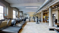 The fitness center at 390 Capitol is a generously proportioned, single-floor facility designed to support a comprehensive workout experience. The space showcases an industrial aesthetic, with exposed ductwork running along the ceiling, painted wooden beam detailing overhead, and arched brick windows lining one side of the room that allow natural light to filter in and create an open, airy atmosphere. The flooring transitions between light wood and modular carpet tiles, defining distinct workout zones throughout the space. The main cardio area is lined with a robust selection of equipment, including treadmills, elliptical trainers, and stationary bikes from brands such as TRUE Fitness, arranged in orderly rows for ease of use. A full-length mirrored wall anchors one end of the room, enhancing the sense of depth and allowing users to monitor their form during exercise. Toward the rear of the facility, a dedicated strength and functional training zone is visible, featuring wall-mounted equipment, exercise balls, and additional resistance machines. The open layout and thoughtful organization of the space make it easy to navigate between cardio and strength training areas, offering a well-rounded fitness environment suitable for a variety of workout routines.