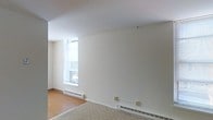 This single-floor space presents a clean, minimalist aesthetic characterized by smooth white walls and a simple, uncluttered layout. The main living area features a combination of hard surface flooring and low-pile carpet, offering a subtle distinction between zones within the open space. A prominent window along one wall allows natural light to filter in, brightening the interior and contributing to an airy atmosphere. The ceilings are a standard height, maintaining a comfortable and proportionate sense of scale. Baseboard heating units run along the lower perimeter of the walls, providing a practical and unobtrusive heating solution. Electrical outlets are positioned at convenient intervals along the walls. The overall design is neutral and functional, with white walls offering a versatile backdrop suitable for a variety of uses. The space is currently vacant, allowing for a clear assessment of its layout and structural features. Its straightforward design and practical amenities make it an adaptable and functional environment.