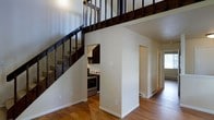 This two-story space is defined by its open, airy layout and thoughtful use of vertical space. A bold staircase with dark metal railings and wood treads serves as a central architectural feature, drawing the eye upward to the second floor, where an open mezzanine-style landing overlooks the main level below. The generous ceiling height enhances the sense of spaciousness throughout the main floor.

The kitchen is efficiently positioned beneath the staircase, making clever use of the available footprint. It features dark cabinetry and stainless steel appliances, including a range, and is tucked neatly into the space without feeling confined. The open sightlines from the kitchen connect to the surrounding living areas, maintaining a cohesive flow throughout the main level.

Warm hardwood flooring runs throughout the main living area, adding both visual warmth and a sense of continuity. The neutral, light-toned walls provide a clean backdrop that complements the darker accents of the staircase and cabinetry. A hallway visible toward the back of the space leads to additional rooms, suggesting a well-organized and functional floor plan. Natural light filters in through a window at the rear, brightening the space naturally.