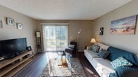 This single-floor unit offers a thoughtfully designed layout with a cohesive neutral color palette and dark hardwood-style flooring throughout the main living areas. The living room serves as the central gathering space, featuring a textured ceiling, warm taupe walls, and a large sliding glass door fitted with vertical blinds that floods the room with natural light and provides direct access to an outdoor patio with greenery views.

Adjacent to the living room, a dedicated dining area accommodates an intimate seating arrangement beneath a ceiling fan with integrated lighting, adding both function and charm. A framed mirror and artwork enhance the welcoming atmosphere of this space.

The galley-style kitchen is efficiently organized with white cabinetry, dark granite-style countertops, a mosaic tile backsplash, and black appliances including a refrigerator, electric range, and over-the-range microwave. Track lighting and wood-style flooring complete the kitchen's practical yet polished look.

The bedroom is a calm retreat featuring plush carpet, soft gray walls, a window with sheer curtains allowing natural light, and ample space for a full suite of furniture. The full bathroom includes a large vanity mirror, dark countertop, a tub-shower combination, and bright overhead lighting.