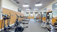 This fitness center in Jacksonville, FL (32257) is a single-floor facility designed to support a comprehensive workout experience. The space features a bold two-tone color scheme, with warm amber-orange accent walls contrasted against crisp white upper walls, creating an energizing atmosphere throughout. Recessed panel lighting in a drop ceiling ensures the room is consistently and evenly illuminated, complementing the natural light that filters in through a row of windows along one wall, offering views of the surrounding greenery.

The cardio section is prominently featured along one side of the facility, housing a lineup of Spirit-brand treadmills and SportsArt elliptical cross-trainers, providing ample options for aerobic exercise. On the opposite side, a full wall of floor-to-ceiling mirrors visually expands the space and allows for form monitoring during workouts. The strength training area includes multi-station cable and weight machines, a rowing machine, and a stability ball for functional fitness. A weight bench is also available for free weight exercises.

Wall-mounted flat-screen TVs are positioned throughout the room, offering entertainment during cardio sessions. The flooring is durable rubber gym matting, suited to high-traffic fitness use. A small adjoining lounge area with seating is accessible through an interior doorway, providing a convenient space to rest before or after workouts.