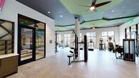 Located at 1515 Rollick Rd in Cary, NC, this well-appointed fitness center spans a generous single floor and is divided into two distinct exercise rooms connected by hallways, offering a comprehensive workout environment for all fitness levels.

The main exercise room is a spacious, open-plan area characterized by striking architectural ceilings with dark charcoal panels accented by sage green geometric shapes, complemented by large ceiling fans and recessed lighting. Tall, black-framed windows line the walls, flooding the space with natural light and providing views of the surrounding community. White structural columns punctuate the floor plan, adding a clean, modern aesthetic. The room is equipped with an extensive array of strength-training machines, cable systems, free weights, benches, rowing machines, and a variety of cardio equipment including treadmills, ellipticals, and stationary bikes. Black-framed glass partition walls and doors seamlessly connect the rooms while maintaining visual openness.

The secondary exercise room serves as a dedicated studio space, featuring a dark ceiling with pendant lighting, a wall-length mirror with a ballet barre, yoga mats, spin bikes, stability balls, and a mounted display screen. A topographic-inspired accent wall adds a distinctive design touch, creating an energizing yet focused atmosphere ideal for cycling classes, yoga, and functional training. Together, both rooms deliver a versatile, modern fitness experience.