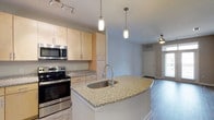 This single-floor, one-bedroom residence is designed with a modern, open-concept layout that promotes a seamless flow between the kitchen and living areas. The kitchen is a standout feature, showcasing a large central island topped with speckled granite countertops and an undermount sink with a sleek gooseneck faucet. Pendant lighting hangs above the island, providing both task illumination and a stylish focal point. Light maple cabinetry lines the kitchen walls, offering generous storage, while stainless steel appliances — including a range and over-the-range microwave — add a polished, contemporary feel. Track lighting along the ceiling further enhances the workspace. The kitchen transitions naturally into the living area, which is finished with wood-style flooring and served by a ceiling fan. Natural light filters in through windows, contributing to a bright and airy atmosphere throughout. The neutral color palette of soft grays and whites lends the space a clean, versatile aesthetic that is easy to personalize. Overall, the residence combines functional design with contemporary finishes to create a comfortable and well-appointed living environment.