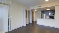 This single-floor space showcases a thoughtfully designed open-concept layout that seamlessly connects the living area and kitchen. Dark hardwood flooring runs throughout, providing a rich contrast against the light, neutral-toned walls and white baseboards, creating a balanced and modern aesthetic.

The kitchen is positioned along one side of the space and features a stainless steel refrigerator, dark cabinetry, and a kitchen island or peninsula with a light-toned countertop, offering both functional workspace and a casual dining or gathering area. The cabinetry's deep hue adds a contemporary accent to the otherwise light interior.

A hallway leads toward additional rooms, with a flush ceiling light guiding the way to what appears to be a main entry door at the far end. A bifold closet door is visible along the living area wall, providing convenient storage without disrupting the open feel of the space.

Overhead, flush-mounted lighting and ceiling ventilation grilles reflect a clean, practical approach to the interior design. The overall ambiance is modern and minimalist, with a neutral palette and quality finishes that lend the space a polished, move-in-ready character. The open sightlines from the living area to the kitchen contribute to an airy and connected feel throughout.