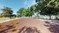 Located at 6710 Wahoo Drive in Myrtle Beach, South Carolina, this outdoor dog park offers a dedicated, fully enclosed space for pets to roam and play freely. The park is secured by a black chain-link fence that runs the perimeter, providing a safe environment for dogs of all sizes. The ground is covered with a generous layer of wood chip mulch, offering a comfortable and low-maintenance surface throughout the enclosure. Mature trees — including pines, oaks, and other native species — are scattered across and around the park, providing ample natural shade during sunny South Carolina days. The tree canopy creates a cool, inviting atmosphere that makes the space enjoyable in warm weather. Views from within the park extend to a nearby pond, adding a pleasant natural backdrop to the setting. Additional glimpses of the surrounding residential neighborhood are visible beyond the fence line. A utility shed is located just outside the fenced boundary, and benches are positioned within the park, offering a place for owners to relax while their pets enjoy the space. The overall setting is open, airy, and well-suited for outdoor recreation, set beneath bright blue skies characteristic of the Myrtle Beach coastal region.
