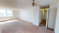 This single-floor space presents a spacious and open living area designed with simplicity and functionality in mind. The main living area is generously proportioned, featuring wall-to-wall plush carpeting in a neutral beige tone that extends throughout, creating a warm and cohesive look underfoot. Crisp white walls provide a clean, bright backdrop, while white baseboard trim adds a subtle finishing detail throughout.

A standout feature of the living area is the large window dressed with vertical blinds, allowing an abundance of natural light to flood the interior and offering views of the surrounding outdoors. Beneath the window sits a baseboard heating unit, providing an efficient heating solution. A ceiling fan with an integrated light fixture is centrally positioned, offering both air circulation and ambient lighting.

The layout flows naturally toward a hallway visible from the main living space, suggesting access to additional rooms beyond. The textured ceiling adds a classic touch to the overall design. The space presents a neutral, move-in-ready environment that offers flexibility for a variety of furniture arrangements and personal styling choices.