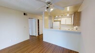 This single-floor, one-bedroom, one-bathroom unit offers an efficient and functional layout designed to maximize the available living space. The open-concept living and kitchen area forms the heart of the unit, where warm hardwood-style flooring runs throughout, adding a cohesive and inviting feel underfoot.

The kitchen is positioned along one side of the space, defined by a peninsula counter that doubles as a breakfast bar or prep area. White cabinetry lines the upper portion of the kitchen, providing ample storage, while a full-size refrigerator and stovetop round out the appliances. Track lighting overhead ensures the kitchen remains well-lit and functional.

The living area benefits from a ceiling fan with integrated lighting, offering both comfort and illumination. A textured ceiling adds subtle visual interest throughout the space.

Toward the rear of the unit, a hallway leads to the bedroom and a built-in shelving unit that provides convenient open storage. Additional closet space is visible, contributing to the unit's overall practicality. The neutral wall tones and white trim create a clean, bright atmosphere throughout the home.