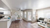 This single-floor residence offers a thoughtfully designed open-concept layout that seamlessly connects the living, dining, and kitchen areas. The living room is bathed in natural light from multiple windows dressed with horizontal blinds and sheer curtains, complemented by wood-look plank flooring and a ceiling fan overhead. A dining nook with a round table and white chairs sits adjacent to the living space, with a round gold-framed mirror adding a decorative touch to the neutral-toned walls. Direct access to an outdoor area is provided through glass-paned doors.

The kitchen is well-equipped with white cabinetry, stainless steel appliances including a range, microwave, and dishwasher, along with a laminate countertop peninsula that doubles as a breakfast bar. A skylight above brightens the workspace throughout the day.

The bedroom is generously proportioned, featuring plush carpet flooring, a large window with outdoor views, and access to a walk-in closet. The two bathrooms are finished with vessel sinks, wood-tone laminate countertops, large framed mirrors, and shower-tub combinations, presenting a clean and modern aesthetic throughout. The overall ambiance is bright, neutral, and move-in ready.
