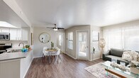 This single-floor residence offers a thoughtfully designed open-concept layout that seamlessly connects the living, dining, and kitchen areas. The living room is bathed in natural light from multiple windows dressed with horizontal blinds and sheer curtains, complemented by wood-look plank flooring and a ceiling fan overhead. A dining nook with a round table and white chairs sits adjacent to the living space, with a round gold-framed mirror adding a decorative touch to the neutral-toned walls. Direct access to an outdoor area is provided through glass-paned doors.

The kitchen is well-equipped with white cabinetry, stainless steel appliances including a range, microwave, and dishwasher, along with a laminate countertop peninsula that doubles as a breakfast bar. A skylight above brightens the workspace throughout the day.

The bedroom is generously proportioned, featuring plush carpet flooring, a large window with outdoor views, and access to a walk-in closet. The two bathrooms are finished with vessel sinks, wood-tone laminate countertops, large framed mirrors, and shower-tub combinations, presenting a clean and modern aesthetic throughout. The overall ambiance is bright, neutral, and move-in ready.