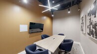 Located at 2901 Connecticut Avenue Northwest in Washington, DC, this professional conference room offers a thoughtfully designed meeting space ideal for small group collaboration. The room centers around a light wood rectangular conference table paired with four upholstered navy blue swivel chairs, creating a polished yet comfortable setting for meetings and presentations.

The space features a striking dual-tone wall design, with a warm amber accent wall serving as the backdrop for a wall-mounted flat-screen display, complemented by a whiteboard panel for collaborative work. The opposite walls are finished in a soft neutral gray, adorned with two bold black-and-white abstract art pieces that lend a sophisticated, creative touch to the interior.

Overhead, an exposed dark ceiling with industrial ductwork adds architectural character, while a geometric star-shaped linear LED chandelier provides focused, contemporary illumination. A small window allows a touch of natural light into the space. Textured carpet flooring grounds the room with a practical, professional feel.

A glazed doorway connects the conference room to a larger adjacent common area, offering visibility into a broader collaborative workspace with bar-height seating and additional lighting. The overall ambiance strikes a balance between modern professionalism and creative energy, making it a functional and visually engaging environment for focused meetings.