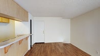 This single-floor space offers a straightforward and functional layout designed for practical everyday living. The main living area features warm, medium-toned hardwood-style laminate flooring that extends throughout, adding a sense of warmth and continuity to the space. Crisp white walls and a textured popcorn ceiling create a clean, neutral backdrop that is easy to personalize.

A notable feature along one wall is a built-in wood-toned cabinetry unit, providing convenient storage and a countertop surface that adds both utility and a touch of natural warmth to the interior. The cabinetry's light wood finish complements the richer tones of the flooring, creating a cohesive aesthetic throughout the space.

A solid white entry door with brass hardware serves as the main point of entry, contributing to the unit's clean and simple design language. The open wall space throughout the living area offers flexibility for furniture arrangement and personalization.

Overall, the space presents a no-frills, move-in-ready environment with practical storage solutions and durable finishes, making it well-suited for those seeking a functional and low-maintenance living space.
