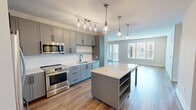 Located on Roland Clarke Place in Reston, VA, this 2-bedroom, 1-bathroom residence offers a thoughtfully designed single-level layout with contemporary finishes throughout. The open-concept kitchen and living area serves as the heart of the home, featuring gray shaker-style cabinetry, white subway tile backsplash, stainless steel appliances, and a light stone countertop island with pendant lighting overhead. Warm hardwood-style flooring extends through the main living areas, complementing the neutral color palette.

The living room is bright and spacious, benefiting from large windows and a glass-panel door that opens to a private balcony with neighborhood views. A second room adjacent to the living area, illuminated by a decorative pendant fixture, offers flexible use as a dining space or home office.

The primary bedroom is generously sized with corner windows on two walls, filling the carpeted room with natural light and views of the surrounding Reston community. A second carpeted bedroom provides a similarly comfortable retreat. Both bedrooms feature neutral tones and clean, modern detailing.

The bathroom is well-appointed with a gray vanity, stone-look countertop, hardwood-style flooring, and a tub-shower combination clad in large-format dark tile with matte black fixtures and built-in shelving niches.