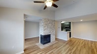 This single-floor space is designed with an open-concept layout that seamlessly connects the living area to the kitchen. The focal point of the living room is a floor-to-ceiling stacked-stone fireplace, framed in a dark surround and set on a tiled hearth, adding both texture and warmth to the space. Light hardwood-style flooring runs throughout, lending a clean, cohesive look to the interior. A ceiling fan overhead provides both function and a subtle design accent, while the elevated ceiling height adds a sense of openness and airiness to the room. Neutral gray walls provide a versatile backdrop that complements the natural tones of the stone and flooring. The kitchen is accessible via a passthrough opening, offering a semi-open connection to the living space. It features dark cabinetry with a sleek finish, stainless steel appliances including a microwave and range, and a light-colored countertop that brightens the area. The overall ambiance of the space is modern and inviting, balancing contemporary finishes with warm, natural materials throughout.