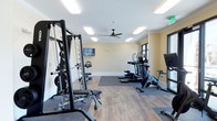 Located at Indigo Square in Mount Pleasant, South Carolina, this fitness center offers a clean, modern environment designed to support a comprehensive workout experience. The single-floor facility is thoughtfully laid out to accommodate a variety of exercise routines. The space features a combination of durable hardwood-style flooring and carpeted zones, clearly delineating areas for cardio and strength training. On the cardio side, a row of elliptical machines and upright exercise bikes lines the room, each equipped with individual display screens for an engaging workout. A multi-station cable and weight machine anchors the strength training area, complemented by additional free weight and resistance equipment. The room is bathed in bright, even light provided by recessed ceiling panels, while a ceiling fan promotes comfortable air circulation throughout sessions. Soft yellow walls contribute to an energizing yet welcoming ambiance. A wall-mounted flat-screen television adds an entertainment option for those looking to stay engaged during longer workouts. Dark-trimmed windows along one wall invite natural light into the space. Crown molding adds a refined architectural touch, elevating the overall finish of this well-appointed community fitness facility.