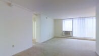 This single-floor space presents a clean and minimalist layout characterized by an open main living area with soft, neutral-toned wall-to-wall carpeting that extends throughout. The room is finished with crisp white walls and a smooth white ceiling, contributing to a bright and airy atmosphere. A large window fitted with vertical blinds dominates one wall, allowing generous amounts of natural light to fill the space. Beneath the window sits a wall-mounted air conditioning unit, complemented by a baseboard heating unit along the perimeter, ensuring year-round climate control. A partial interior wall partially divides the space, adding subtle definition without fully enclosing the layout. A doorway visible toward the rear leads to an adjoining room or corridor, suggesting additional space beyond the main living area. The overall design is straightforward and functional, with neutral finishes that provide a versatile canvas suitable for a variety of furnishing arrangements and interior styles. The absence of decorative elements keeps the focus on the spatial qualities of the unit itself.