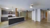 This single-floor loft-style space showcases an open-concept layout that blends industrial and contemporary design elements seamlessly. The polished concrete floors extend throughout, providing a sleek, durable foundation that enhances the modern aesthetic. Structural wooden posts, finished in a warm natural tone, serve as striking architectural focal points that add character and a nod to the building's industrial heritage.

The kitchen is well-appointed with dark wood cabinetry, stainless steel appliances including a refrigerator and range with microwave, and a spacious island featuring a granite-style countertop with a built-in sink. Pendant lighting above the island provides both functional illumination and a refined design touch.

Exposed ductwork and ceiling-mounted light fixtures reinforce the industrial character of the space, while clean white walls and neutral tones keep the atmosphere bright and open. Framed doorways with open shelving and partition elements define separate zones within the open floor plan without sacrificing the sense of spaciousness. The layout offers generous room for flexible use, making it well-suited for both living and entertaining. Overall, the space delivers a cohesive, urban-industrial ambiance with thoughtful modern finishes throughout.