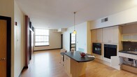 This single-floor space showcases a thoughtfully designed open-plan layout that seamlessly connects the kitchen and living areas. The kitchen serves as the centerpiece, featuring a sleek curved island with a dark countertop, light maple cabinetry, and a built-in wall oven and microwave stack — providing both ample storage and modern functionality. A stylish pendant light hangs above the island, adding a warm focal point to the space.

The flooring throughout is warm-toned hardwood, lending a cohesive and inviting feel across the entire floor plan. White walls and a clean white ceiling enhance the sense of openness and brightness.

One of the most defining features of this space is the generous natural light, courtesy of large, multi-pane industrial-style windows that line the living area. These oversized windows evoke a loft-like character, suggesting a building with architectural heritage. The living area extends comfortably beyond the kitchen, offering a versatile, open space suitable for a variety of furniture arrangements.

Overall, the space balances contemporary finishes with a loft-inspired aesthetic, creating an environment that feels both functional and welcoming — ideal for everyday living and entertaining alike.