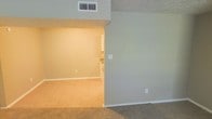 This single-floor residential space offers a straightforward and functional layout designed for comfortable everyday living. The interior is finished with neutral, muted green-gray walls that create a calm and cohesive atmosphere throughout. Soft, wall-to-wall carpeting in a warm beige tone covers the floors, adding comfort underfoot and contributing to the space's understated, livable character.

The main living area flows openly into an adjoining room, which appears to be a kitchen or dining area. This connected layout promotes a sense of continuity and makes the space feel larger than its footprint might suggest. The transition between rooms is defined by a subtle architectural step-down or drop in the ceiling, adding a modest sense of dimension and separation without fully closing off the two areas.

Practical details are evident throughout, including ceiling-mounted HVAC vents for climate control, standard electrical outlets positioned along the baseboards, and clean white trim that frames the walls and transitions neatly between surfaces. The lighting in the adjoining area emits a warm glow, enhancing the inviting quality of the interior. Overall, this space presents a clean, move-in-ready environment with a neutral palette well-suited for personalization.