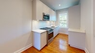 Located at 133 Oxford St in Cambridge, MA, this single-floor unit offers a thoughtfully updated interior with a clean, modern aesthetic. Warm-toned hardwood floors run consistently throughout the space, providing a cohesive and inviting foundation in every room.

The open living area is generously proportioned, bathed in natural light from large double-pane windows that frame views of the surrounding Cambridge neighborhood. Recessed lighting complements the natural light and enhances the bright, airy atmosphere maintained by soft gray-white walls and crisp white trim.

The kitchen is well-appointed with flat-front white cabinetry extending to the ceiling, white quartz countertops, stainless steel appliances including a gas range and built-in microwave, and a single-basin sink positioned beneath a window overlooking greenery. The streamlined layout maximizes functionality without sacrificing style.

The bedrooms are spacious and well-lit, with one room featuring a built-in closet with shelving. Each bedroom benefits from recessed lighting and the same warm hardwood flooring found throughout the unit.

The bathroom is finished with full floor-to-ceiling tile, a glass-enclosed walk-in shower with a handheld fixture, a built-in niche shelf, a soaking tub, and a pedestal sink — presenting a sleek, spa-like environment. The overall design is cohesive, modern, and move-in ready.