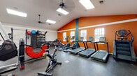 Located at 3770 Southpointe Pkwy in Oxford, Ohio, this single-floor fitness center is designed to support a full range of workout routines in a well-equipped, energizing environment. The space is defined by its striking vaulted ceilings, which create an open and airy atmosphere, complemented by large skylights and multiple windows that flood the interior with natural light. Ceiling fans throughout help maintain a comfortable environment during workouts.

The cardio zone is prominently featured along one wall, lined with treadmills, elliptical machines, a stair climber, and stationary cycling bikes — all positioned near windows to provide natural views during exercise. Bold orange accent walls with teal detailing add a vibrant, motivating energy to this area. The strength training section occupies a separate, generously sized area finished in white and neutral tones, housing a variety of cable machines, weight benches, and selectorized strength equipment arranged for ease of movement and access. A dedicated free weights station includes a full rack of dumbbells and medicine balls.

Entry to the facility is through a glass-paneled door opening into a tiled vestibule area. Durable rubber flooring covers the main workout areas, prioritizing safety and functionality. The overall design balances practicality with an inviting, well-lit atmosphere suited for comprehensive fitness training.