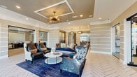 Located at 120 Vintage Foxland Dr in Gallatin, Tennessee, this elegantly appointed community clubhouse spans a generous single floor, offering two distinct living areas and two fully equipped kitchens designed for both social gatherings and everyday comfort. The main lounge greets visitors with herringbone-patterned tile flooring, a striking sputnik chandelier with crystal accents, and a coffered ceiling that adds architectural depth. Built-in shelving with horizontal white trim lines the walls, lending a refined, layered look. A secondary living area features a tile-surround fireplace beneath a wall-mounted TV, a coffered wood-paneled ceiling with integrated lighting, and an arrangement of plush neutral seating. A smaller seating nook near the entrance offers a cozy retreat with natural light pouring through large windows. The two kitchens are thoughtfully designed: one showcases navy cabinetry, white quartz countertops, exposed wood beam ceilings, and a full beverage station with draft taps, while the other offers a bright island with bar seating beneath elegant lantern pendants. Warm neutral tones, curated artwork, and a cohesive blend of modern and transitional design elements create an inviting, upscale ambiance throughout the entire space.