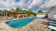 Located at 4950 US-90 W in Mobile, Alabama, this outdoor amenity space centers around a generously sized swimming pool with vivid turquoise water. The pool area is designed for both relaxation and recreation, offering a curved, resort-style layout complemented by a broad paved deck finished in an attractive stone or brick pattern that surrounds the pool on all sides.

A standout architectural feature is the substantial pergola structure along one side of the pool, built with stone columns and a wood-beam overhead framework. This shaded zone is outfitted with multiple teal-colored lounge chairs, providing a comfortable retreat from the Alabama sun. Additional lounge chairs and a patio umbrella are positioned around the pool deck, offering flexible seating options for guests.

A single-story building with vinyl siding and multiple windows sits adjacent to the pool, likely serving as a clubhouse or amenity facility. The entire area is enclosed by a black iron fence, ensuring privacy and security. Towering mature oak and pine trees frame the perimeter, delivering a lush, green backdrop and a sense of seclusion. The space benefits from abundant natural light under open skies, making it an inviting outdoor destination suited for warm Southern weather.